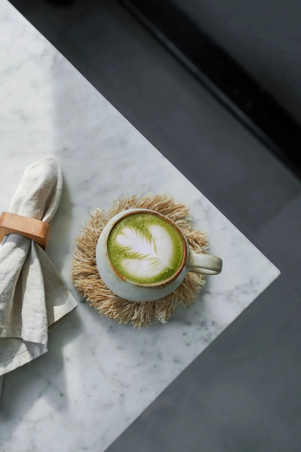 Tasse en céramique blanche pour matcha latte avec motif feuille de thé vert sur mousse, sur dessous de verre rond en raphia naturel à franges solaires